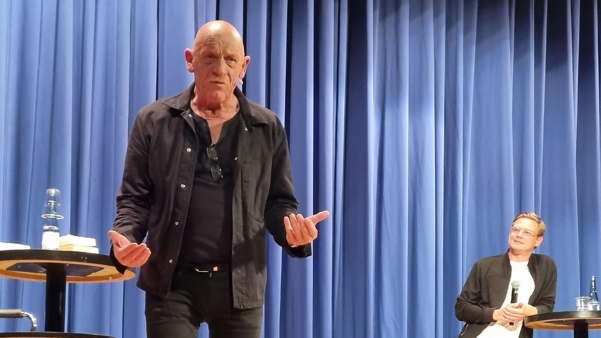 Der Schauspieler und Arzt Joe Bausch steht auf einer Bühne mit geöffneten Händen und gestikuliert in Richtung des Publikums.