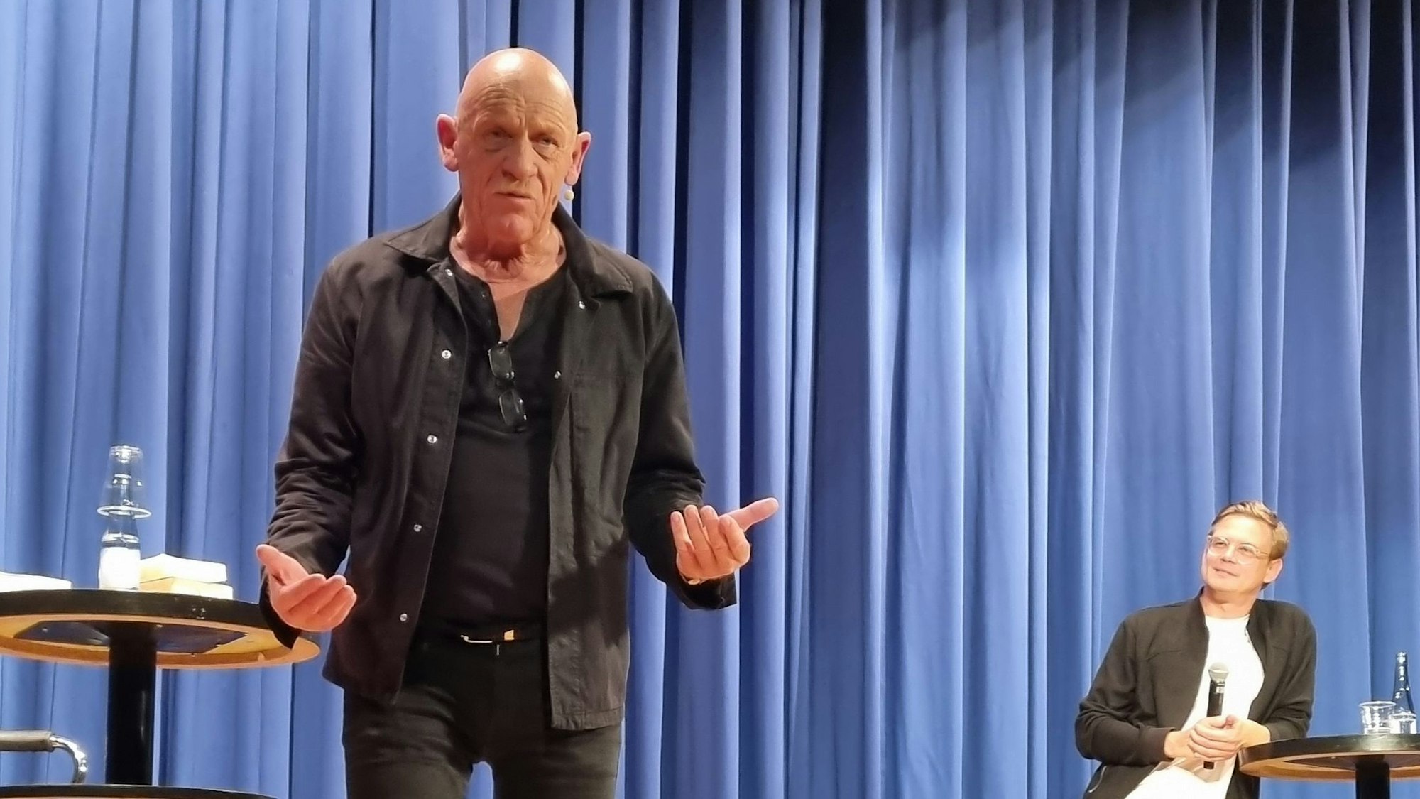 Der Schauspieler und Arzt Joe Bausch steht auf einer Bühne mit geöffneten Händen und gestikuliert in Richtung des Publikums.