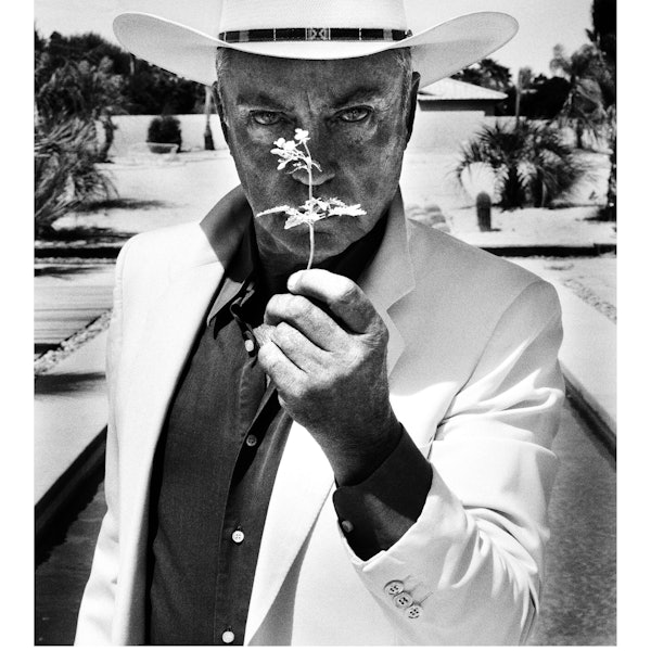 Udo Kier hält eine Blume vor sein Gesicht.