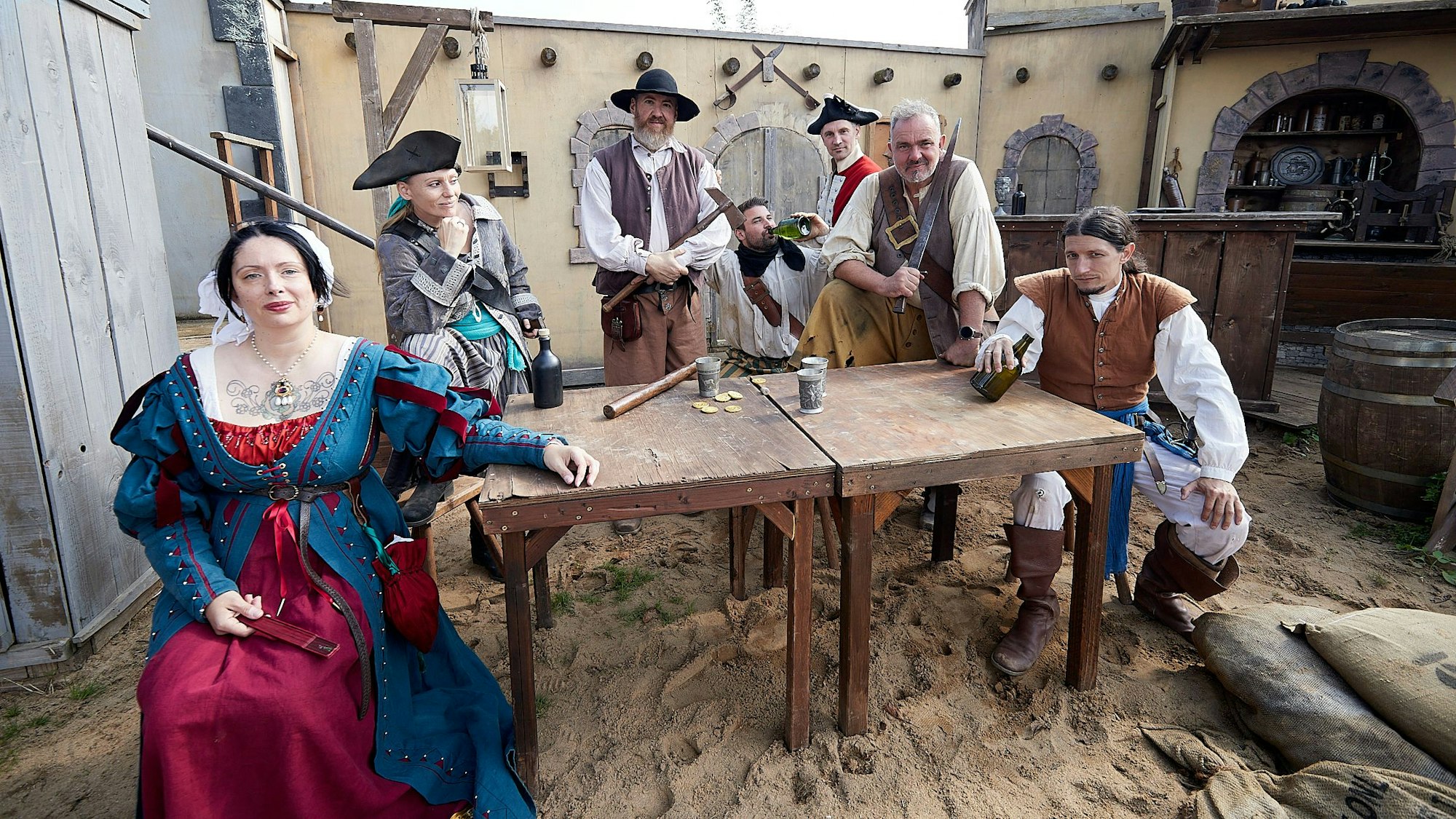 Menschen in Piratenkostümen sitzen an einem Holztisch, der auf sandigem Boden steht. Auf dem Tisch liegen Goldtaler, daneben stehen Trinkbecher.