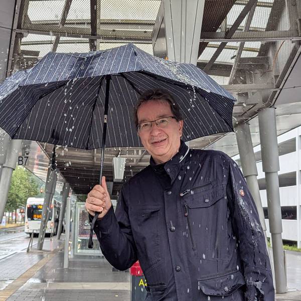 Das Foto zeigt einen Mann mit Regenschirm am Busbahnhof in Hürth Mitte. Auf den Schirm prasselt das Wasser.