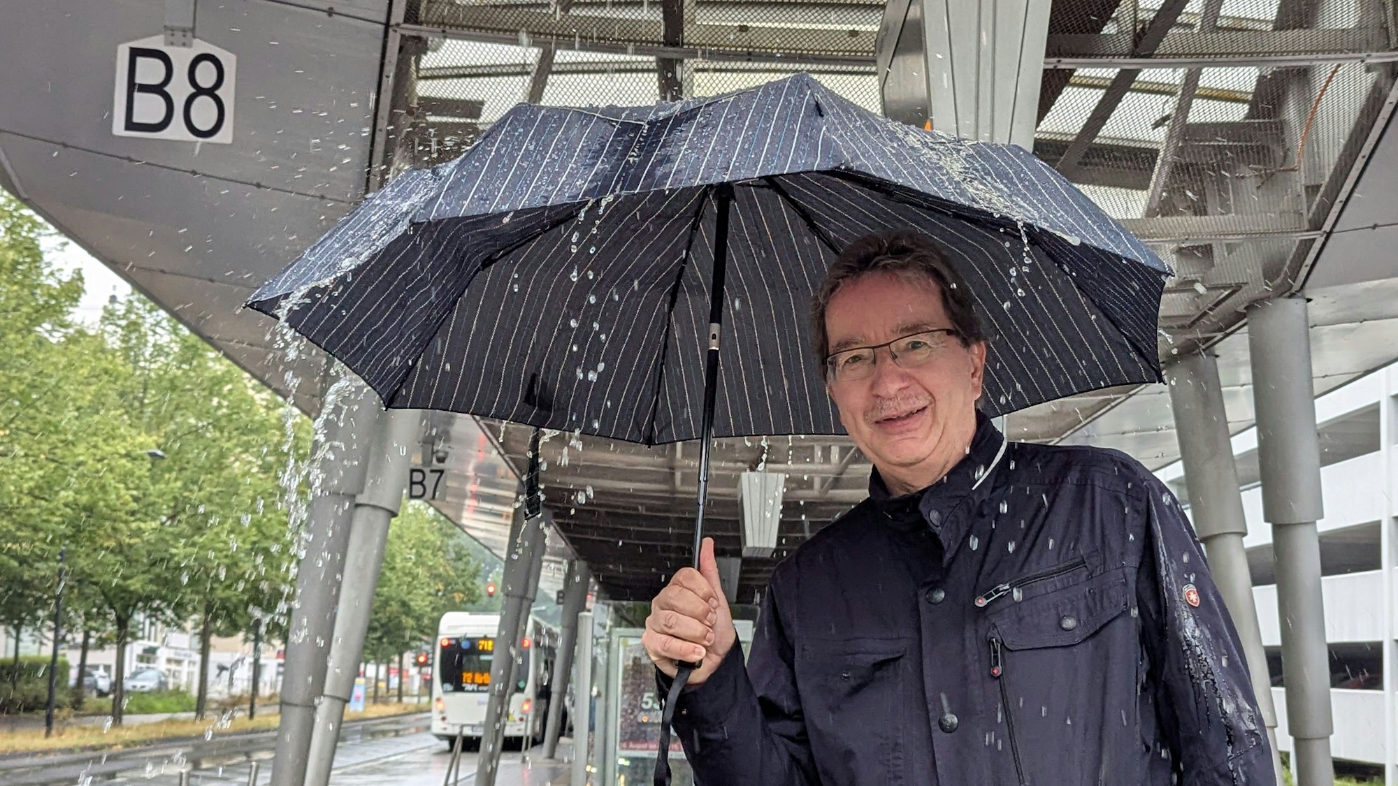 Das Foto zeigt einen Mann mit Regenschirm am Busbahnhof in Hürth Mitte. Auf den Schirm prasselt das Wasser.