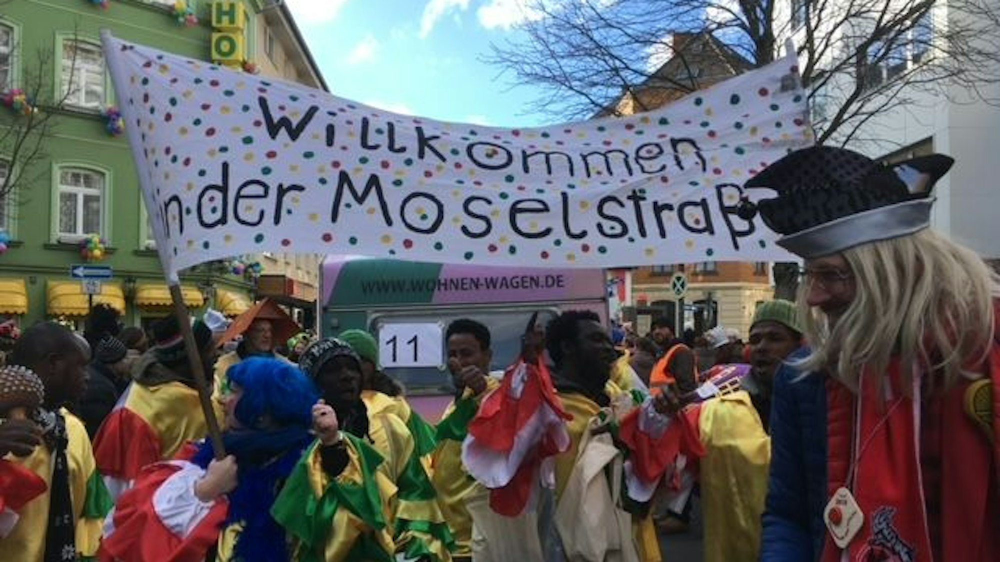 Karnevalistisch verkleidete Menschen stehen rund um ein großes Plakat mit den Lettern Willkommen in der Moselstraße