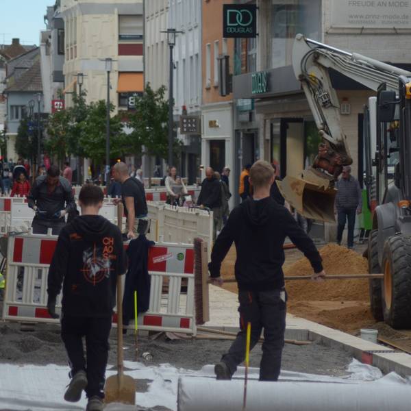 In der Neustraße verlegen Arbeiter hinter Absperrzäunen Pflastersteine. Ein Bagger verteilt Sand, während Passanten sich einen Weg vorbei an der Baustelle bahnen.