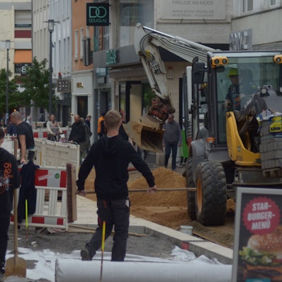 In der Neustraße verlegen Arbeiter hinter Absperrzäunen Pflastersteine. Ein Bagger verteilt Sand, während Passanten sich einen Weg vorbei an der Baustelle bahnen.