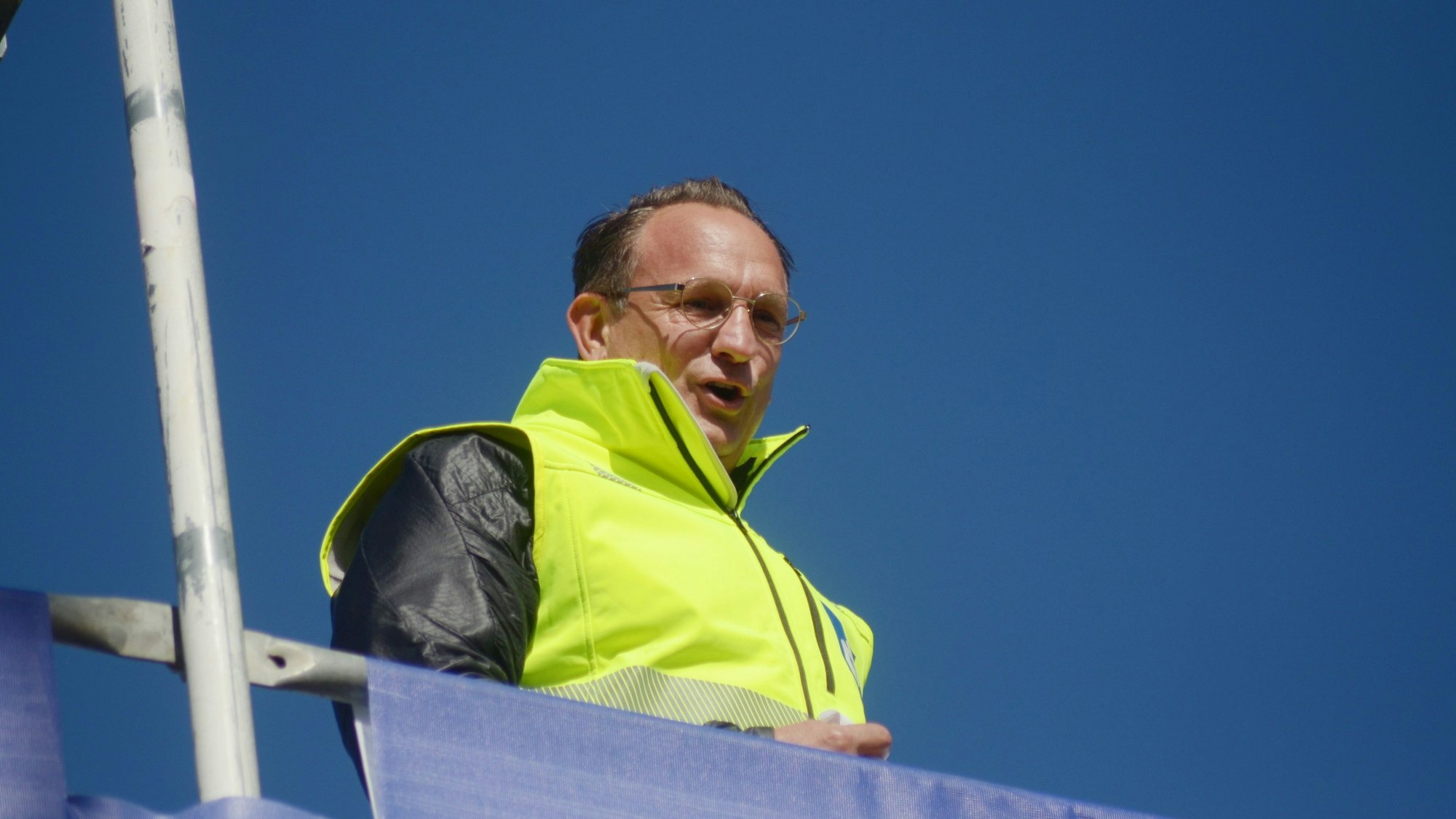 Der Polier, der eine gelbe Warnweste trägt, steht hinter einem Geländer auf dem Kita-Gebäude.