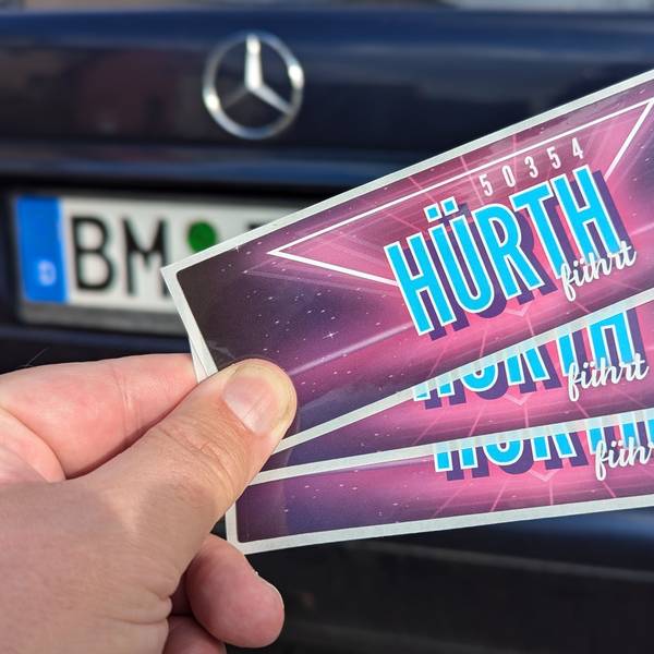 Das Foto zeigt eine Hand, die drei Aufkleber vor einem Auto mit BM-Kennzeichen hält.