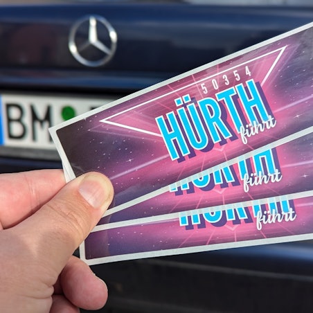 Das Foto zeigt eine Hand, die drei Aufkleber vor einem Auto mit BM-Kennzeichen hält.