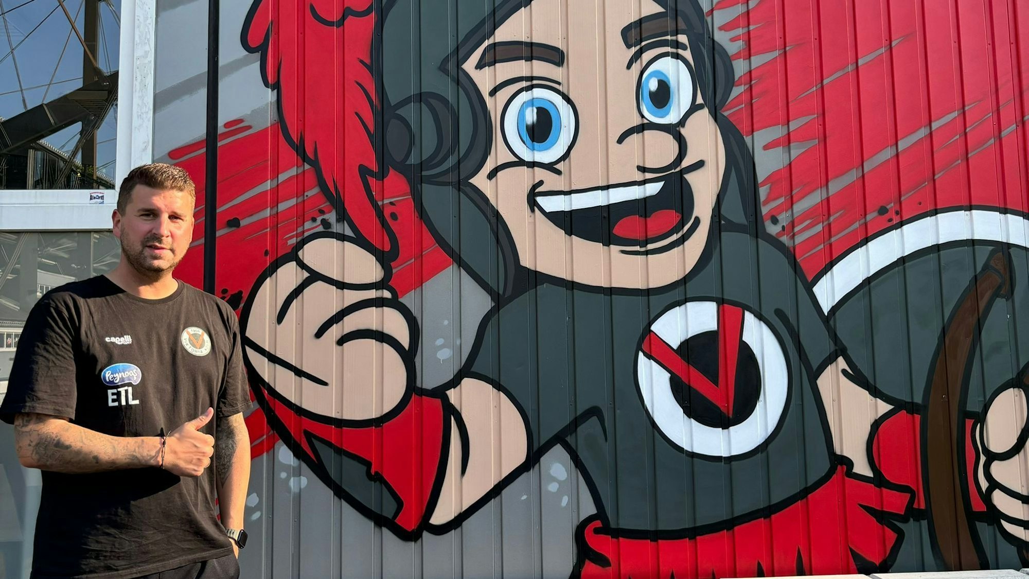 Christian Hammes reckt den Daumen und steht vor einer bemalten Wand bei Viktoria Köln.