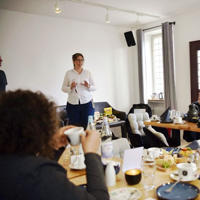 Angelika Eimermacher vom DRK Euskirchen steht in einer weißen Bluse und dunkler Jeans vor einer Gruppe Menschen, die an Tischen sitzen und Kaffee trinken. Auf den Tischen stehen Kaffeetassen, Kuchen und weiteres Geschirr.