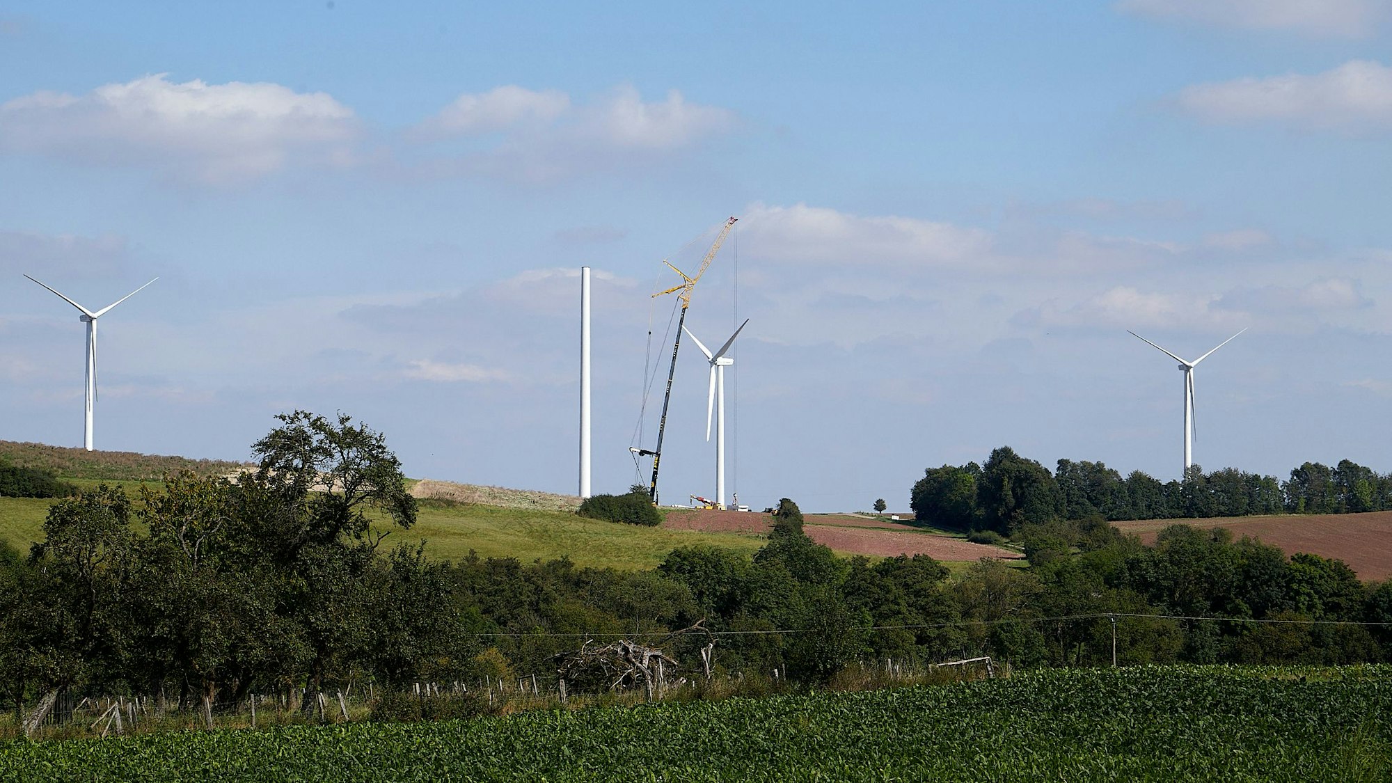 Auf einem Hügel bei Heimbach-Vlatten sind drei Windenergieanlagen zu sehen. Von einer vierten steht der Turm, mithilfe eines Krans wird daran gearbeitet.