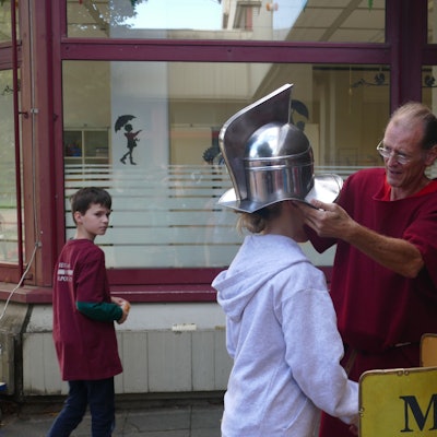 Ein Mann setzt einem Mädchen einen römischen Gladiatorenhelm auf den Kopf.
