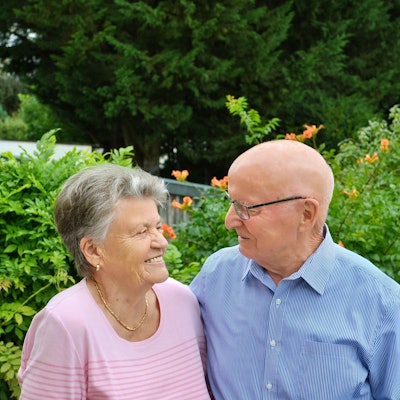 Anna und Heinrich Wagner stehen vor grünen Büschen und schauen sich an. Anna Wagner trägt ein rosafarbenes Oberteil, eine goldene Kette, kleine goldene Ohrringe und kurze graue Haare. Heinrich Wagner trägt ein blaues Hemd und eine Brille, er hat eine Glatze.
