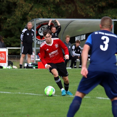 Ein Fußballer hat den Ball am Fuß und setzt zum Dribbling an. Im Vordergrund beobachtet ihn ein Verteidiger.