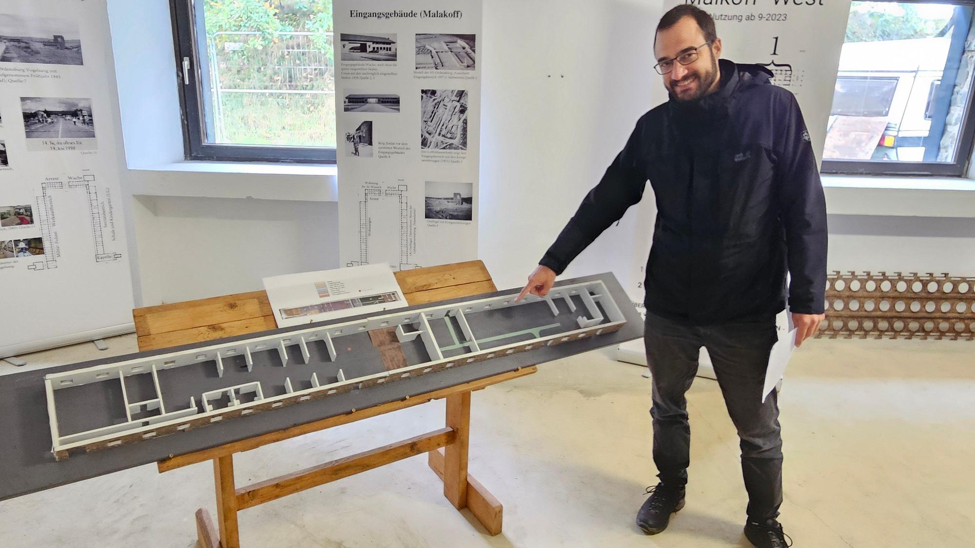 Historiker Julian Weller steht in dunkler Regenjacke und dunkler Jeans neben einem Modell der neuen Ausstellung in Vogelsang. Hinter ihm sind Fenster und Aufsteller zu sehen.