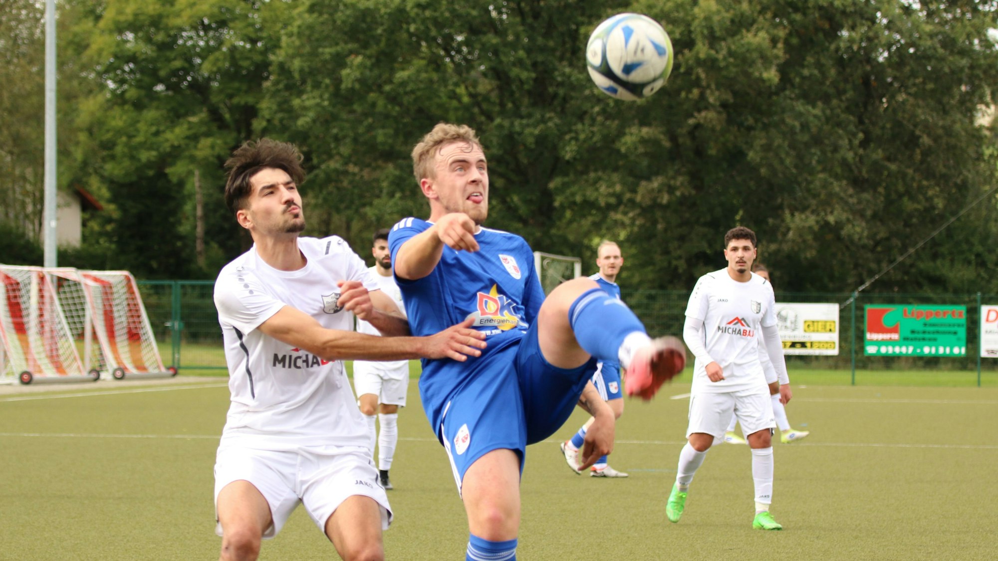 Ein Fußballer schießt einen Ball weg, sein Bein ist fast waagerecht. Ein Gegenspieler hat den Arm an dessen Trikot.