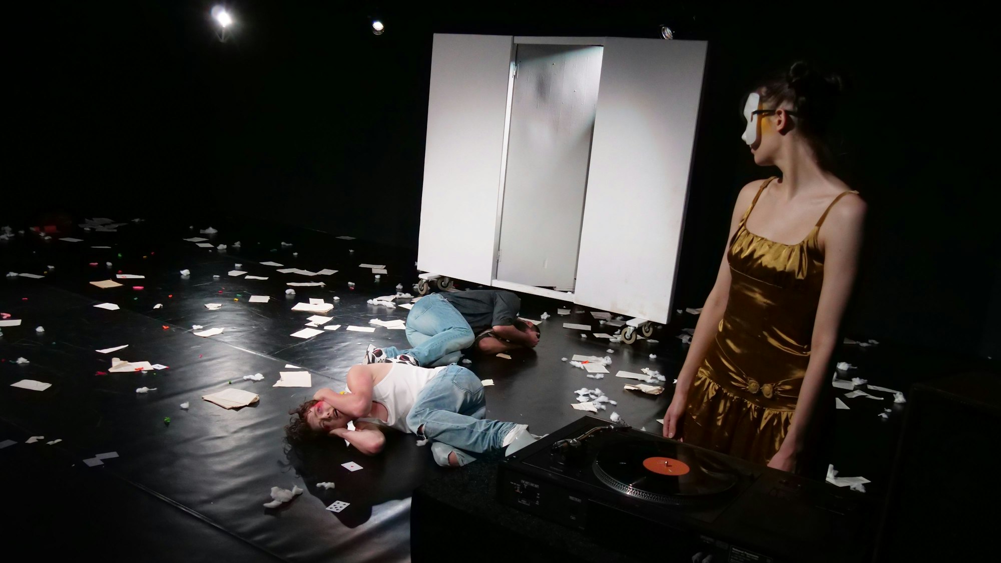 Zwei Schauspielerinnen in einem schwarzen Raum, in dem hinten eine weiße Wand mit Tür ist. Eine Frau trägt eine weiße Maske, eine liegt auf dem Boden.