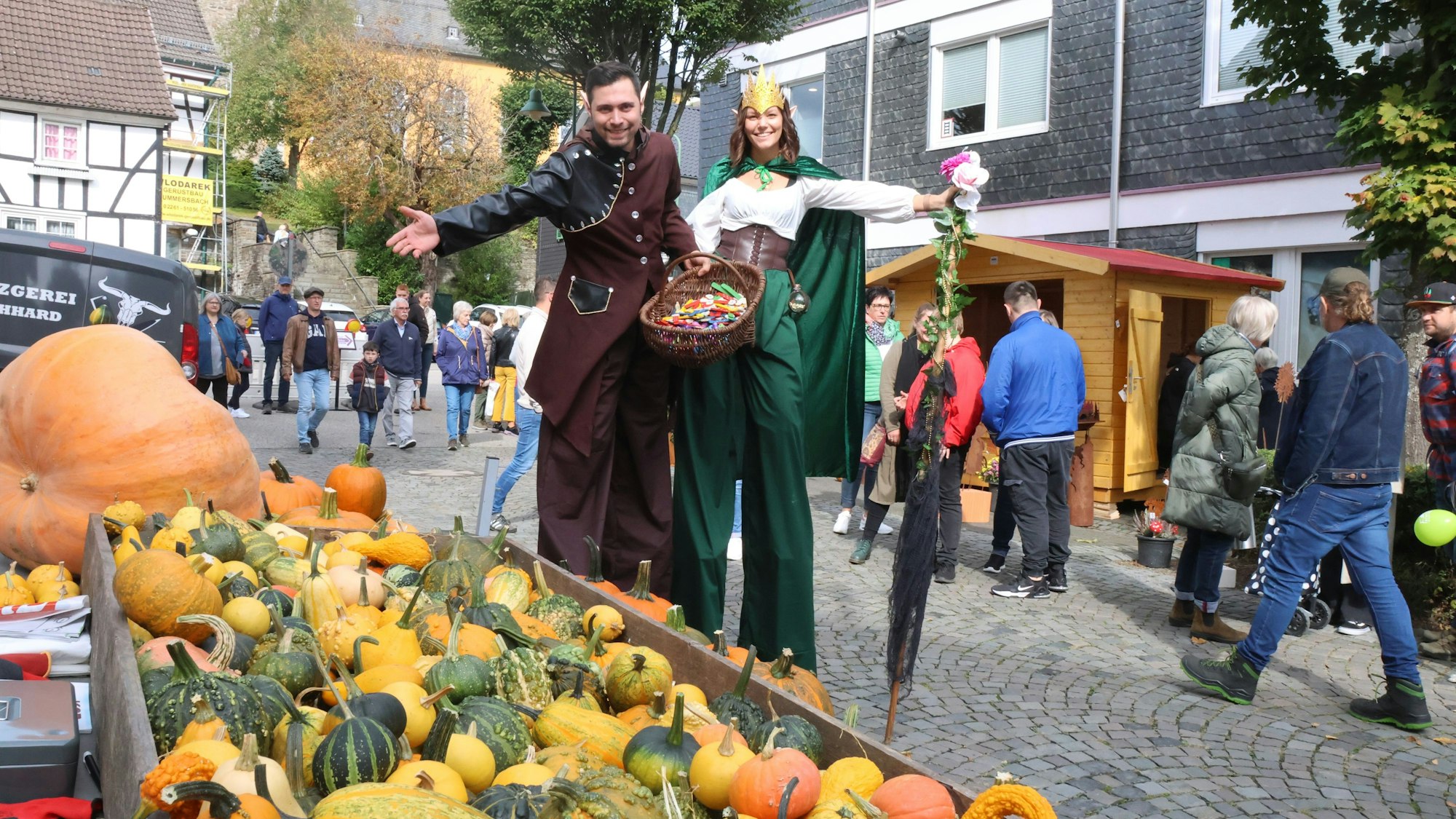 Zwei Personen auf Stelzen und im Waldelfenkostüm verteilen auf einem Erntedankfest Blumen und Süßigkeiten.