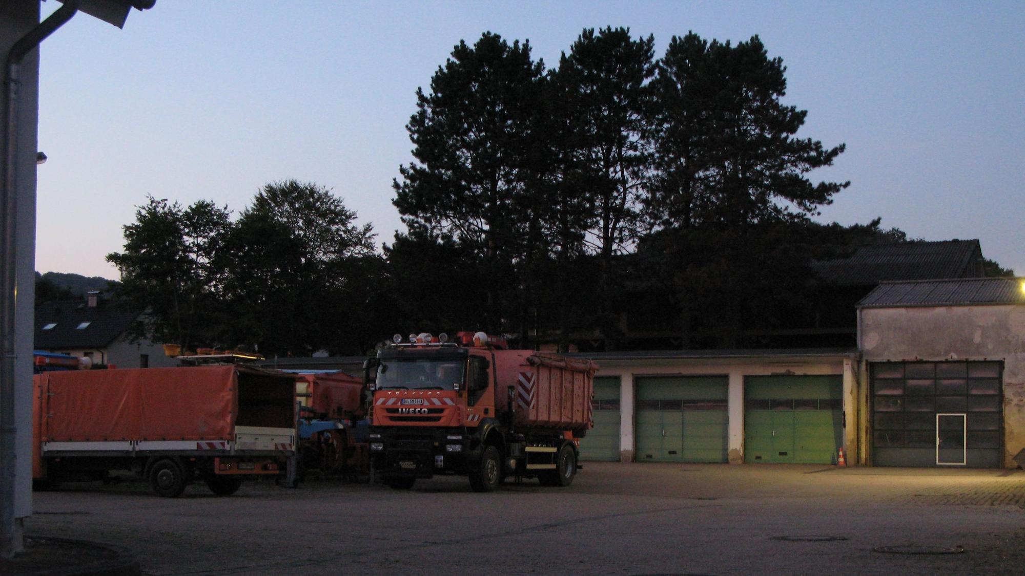 Blick auf einen asphaltierten Hof mit Hallen und orangefarbenen Lastwagen.