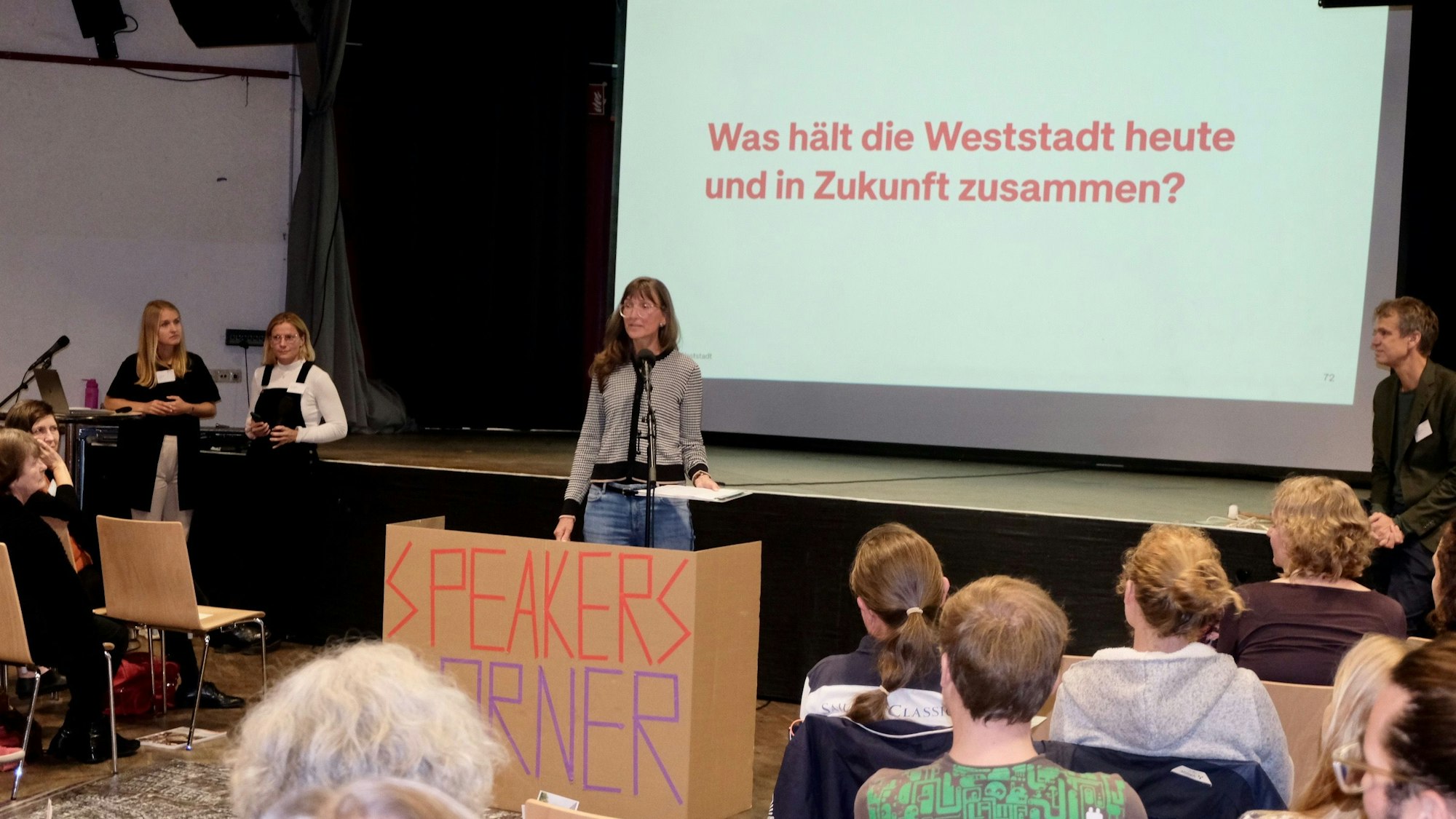 Vor einer Bühne steht eine langhaarige Frau, die auf ein Rednerpult zugeht mit der Aufschrift Speakers Corner.