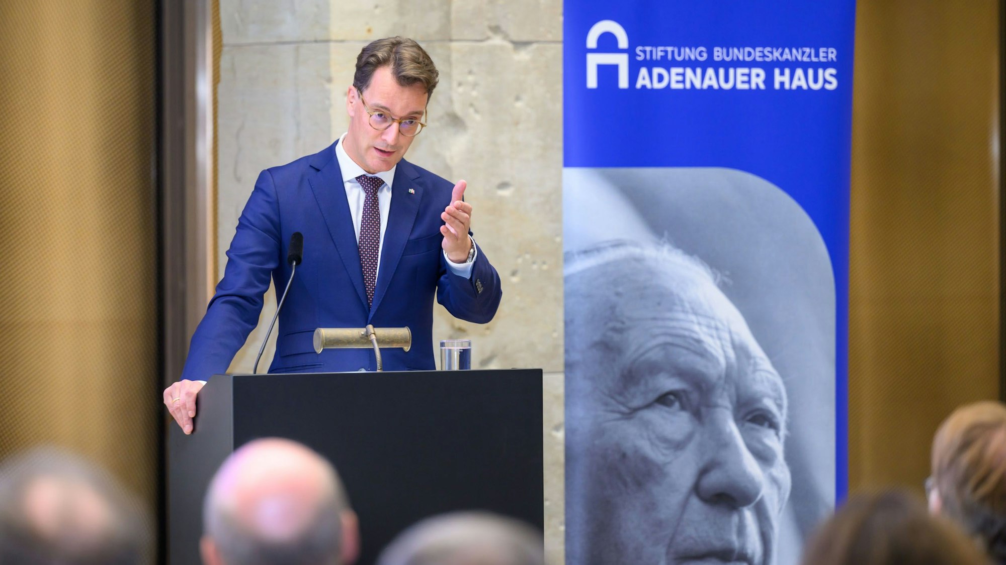 Ein Man steht an einem Pult, daneben befindet sich ein Aufsteller mit einem großen Porträt Konrad Adenauers.