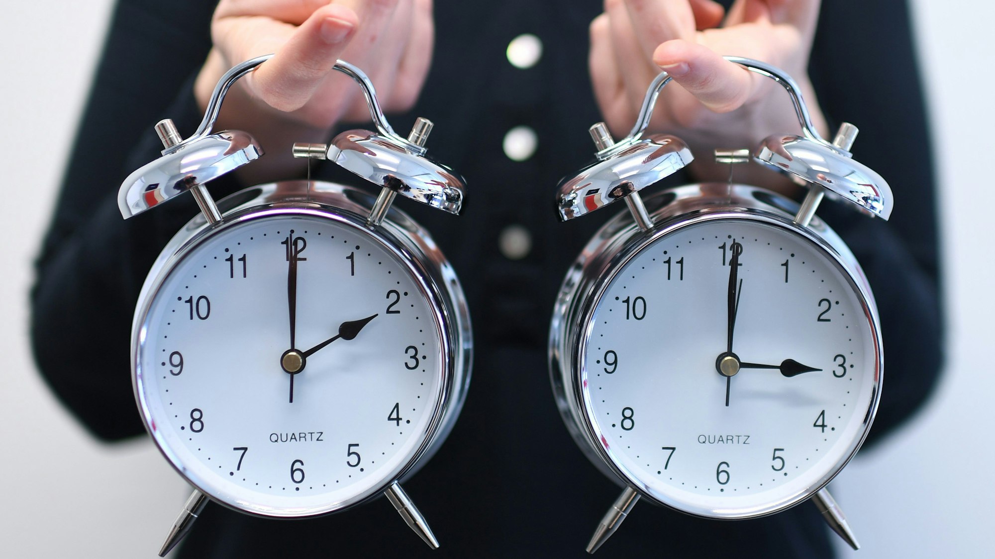Eine Frau hält zwei Wecker in der Hand - der linke zeigt 2 Uhr, im rechten sind die Zeiger auf 3 Uhr gestellt (Archivbild).