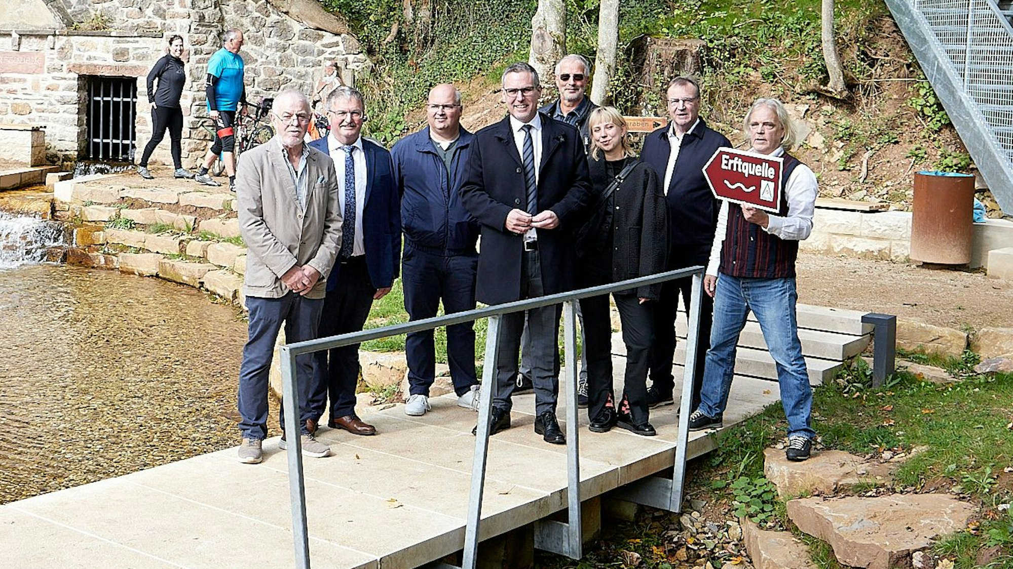 Auf einer neuen Holzbrücke über dem Wasser der Erftquelle stehen acht Personen.