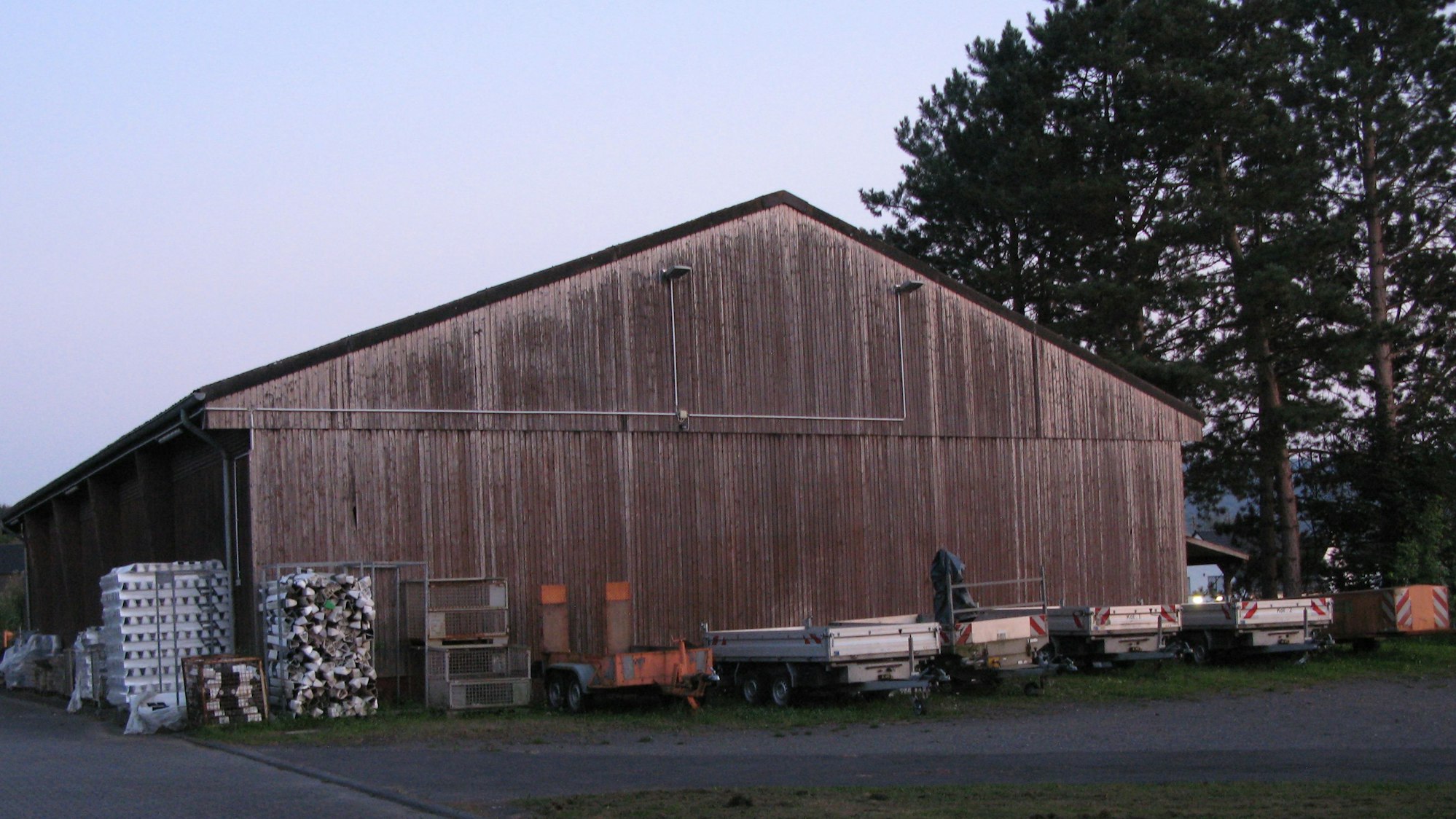 Eine alte Halle mit Satteldach und Holzlattenfassade, vor der einige Anhänger stehen.