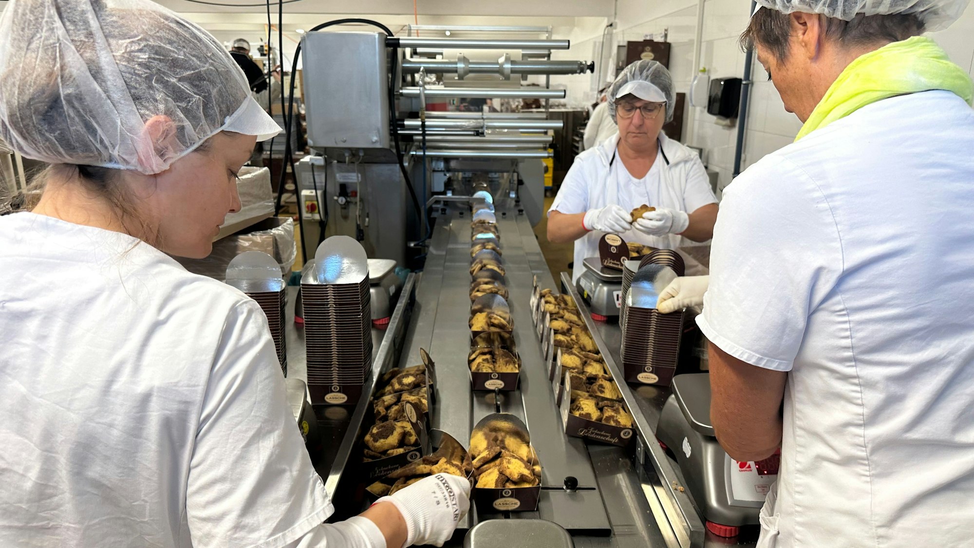 Mitarbeiterinnen wiegen Plätzchen ab und legen sie in die Verkaufsschälchen. Am Ende des Bandes kommt eine Folie über die Verpackung.