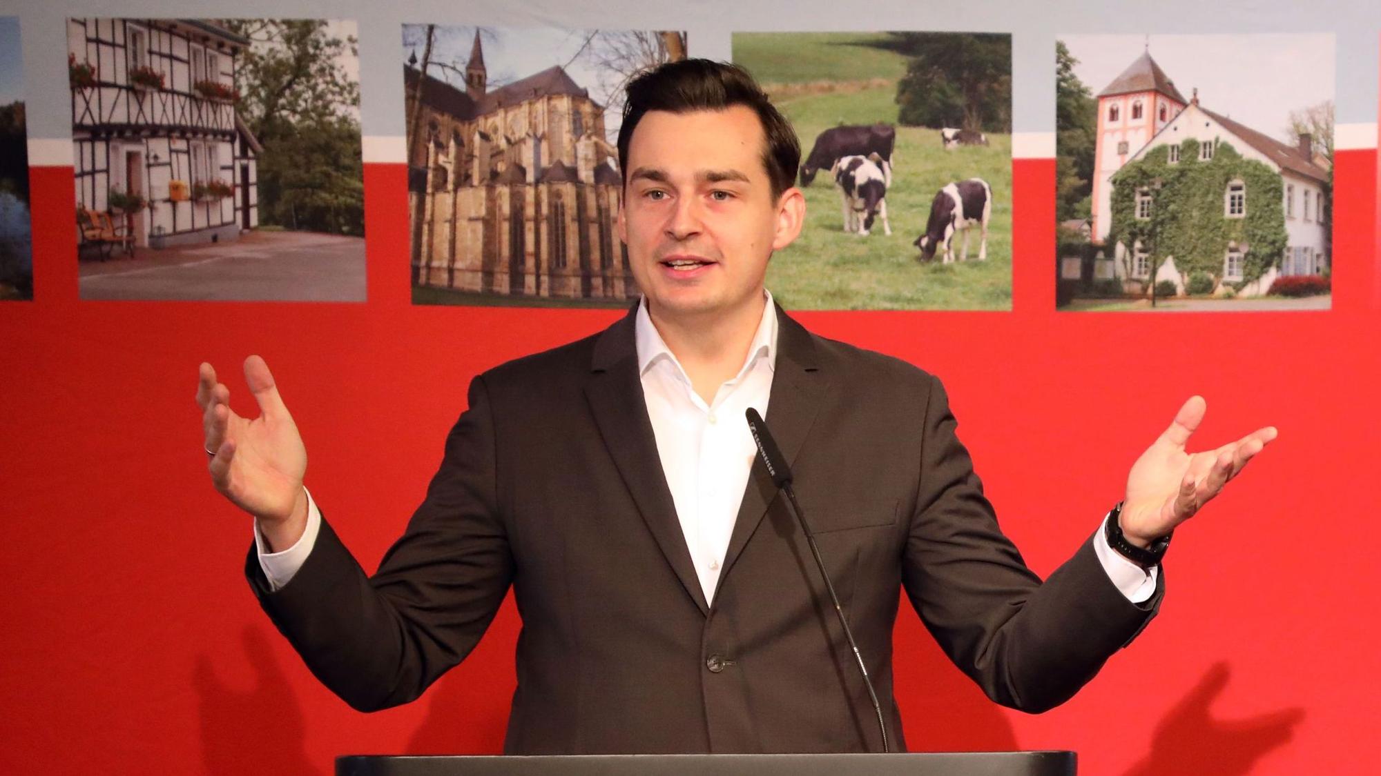 Der Bergisch Gladbacher Bürgermeisterkandidat von SPD und Grünen, Marcel Kreutz (SPD) spricht vor einer roten Wand mit Bildern aus Rhein-Berg.
