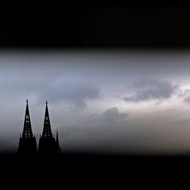 Durch ein Geländer sind die Spitzen des Kölner Doms zu sehen. Im Erzbistum Köln sind die Finanzüberschüsse 2023 eingebrochen.