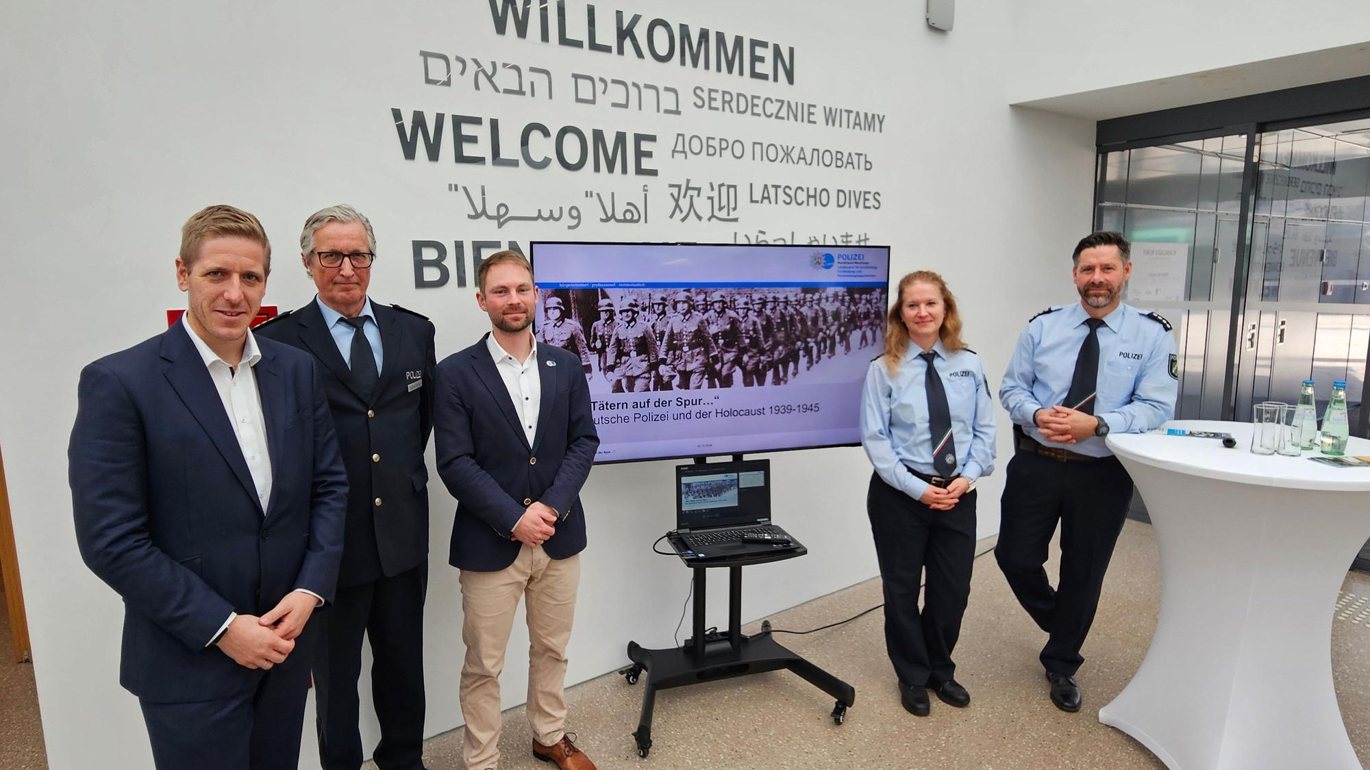Vier Männer und eine Frau stehen nebeneinander und schauen in die Kamera, zwischen ihnen ein Bildschirm mit einem historischen Foto.