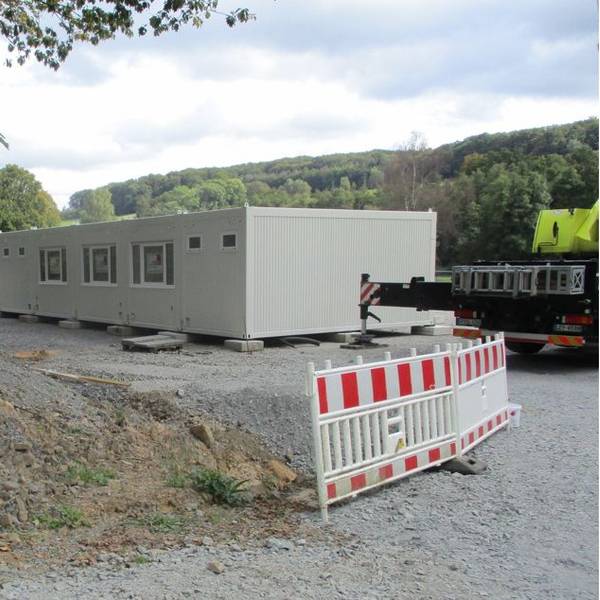 Eine Reihe hellgrauer Wohncontainer auf einem Schotteruntergrund, daneben eine Baustellenabsperrung.