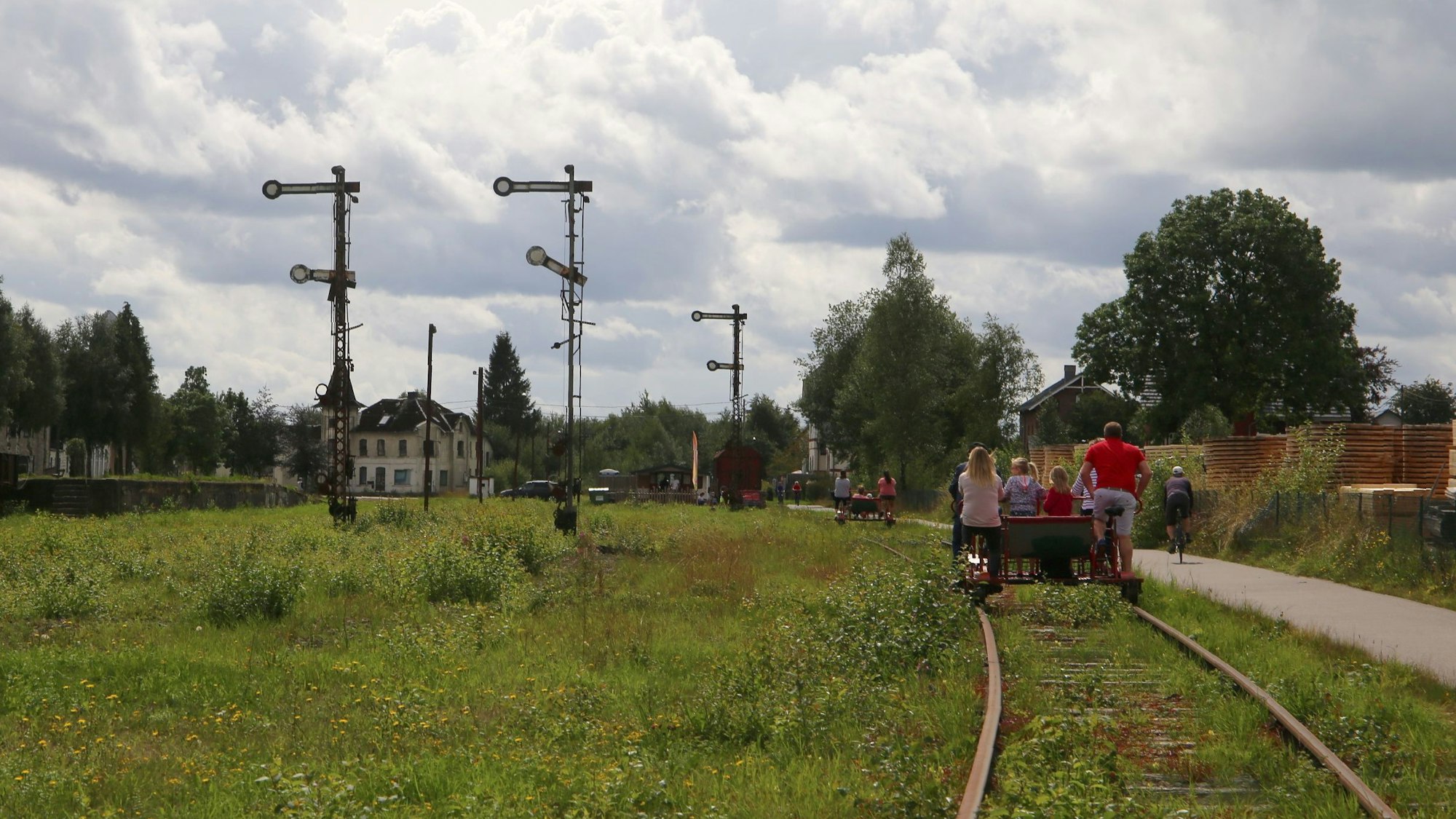 Auf der Trasse der ehemaligen Vennbahn fährt eine Draisine, daneben verläuft ein Radweg. Im Hintergrund stehen alte Bahn-Signalanlagen und ein Bahnhofsgebäude.