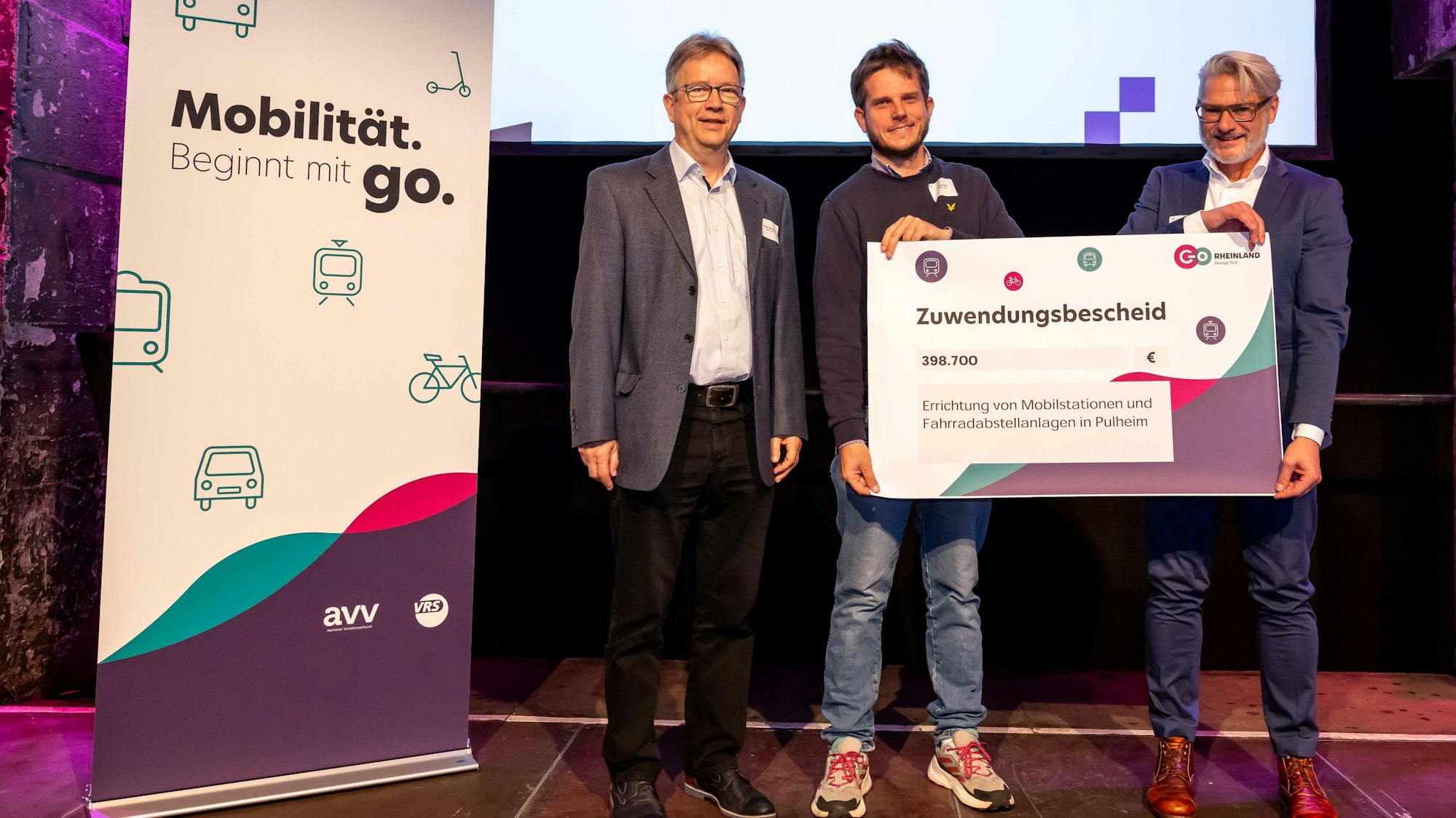 Drei Männer stehen auf einer Bühne, sie lächeln in die Kamera, zwei halten einen großen symbolischen Förderbescheid in den Händen.