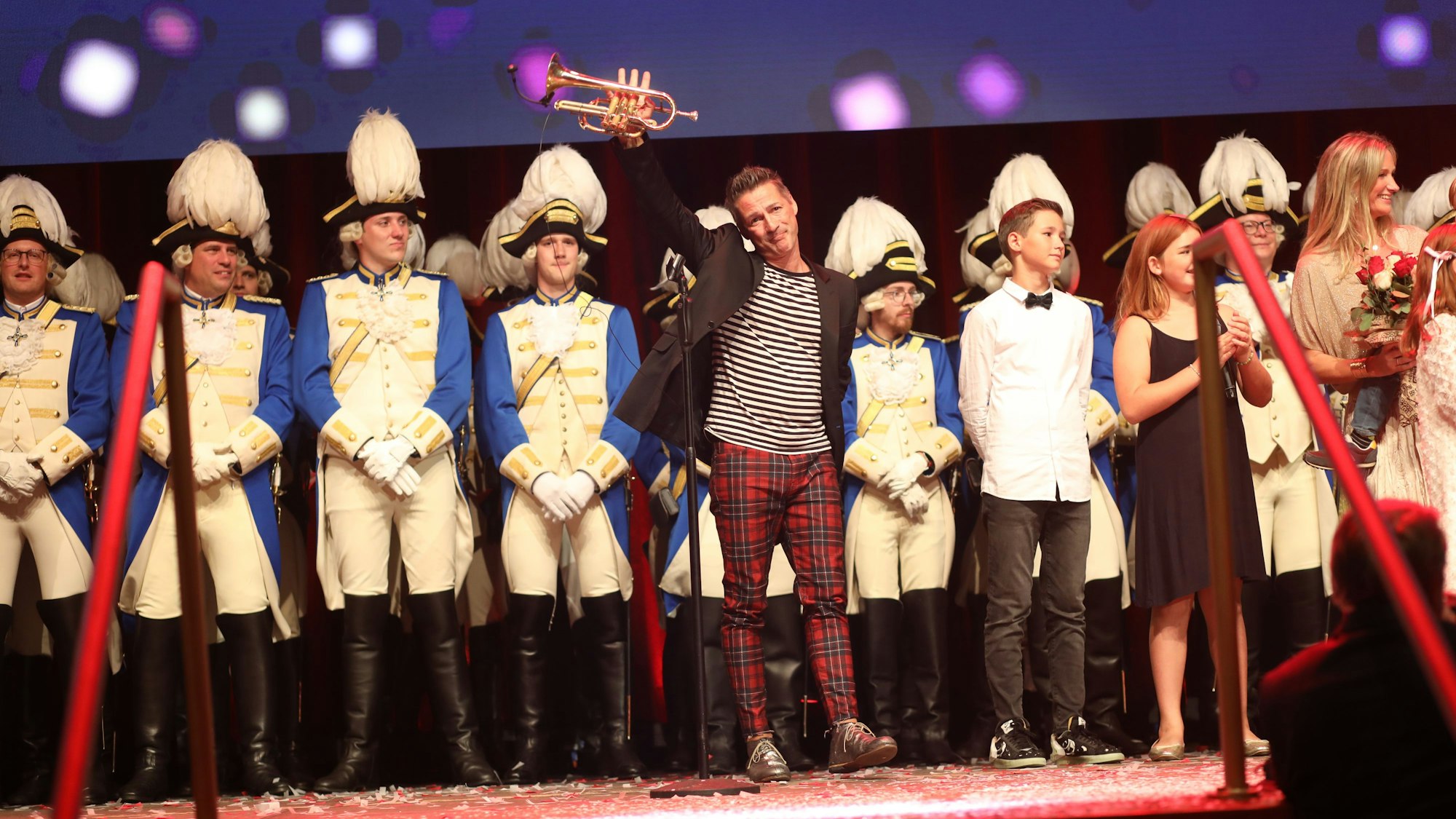 Mann winkt auf Bühne Richtung Publikum, hinter ihm stehen Gardisten.