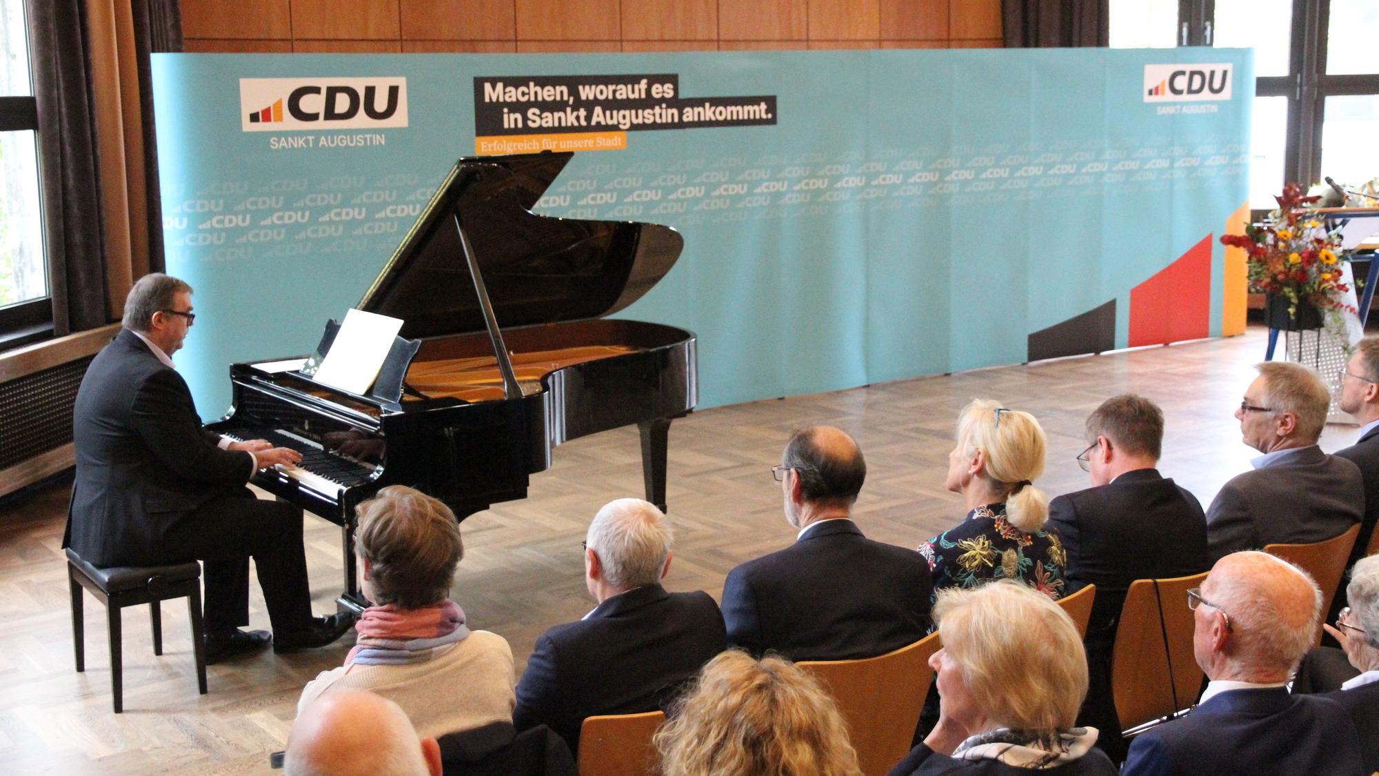Ein Mann an einem Konzertflügel und Zuhörer auf Stühlen in einem Saal