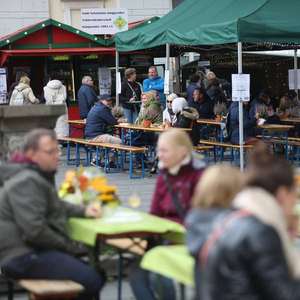 Menschen sitzen an Tischen auf dem Marktplatz, im Hintergrund stehen zwei Weinstände.