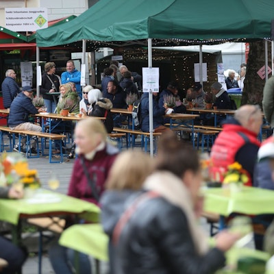 Menschen sitzen an Tischen auf dem Marktplatz, im Hintergrund stehen zwei Weinstände.