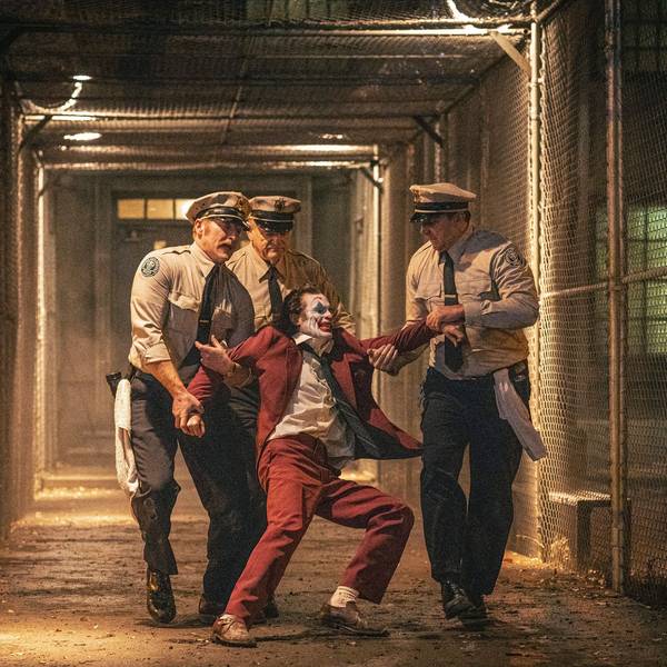 Drei Gefängnisaufseher schleppen einen sich heftig wehrenden Joaquin Phoenix im Joker-Kostüm durch einen Gang zwischen Maschendrahtzäunen zu seiner Zelle zurück.
