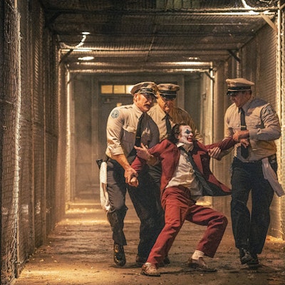 Drei Gefängnisaufseher schleppen einen sich heftig wehrenden Joaquin Phoenix im Joker-Kostüm durch einen Gang zwischen Maschendrahtzäunen zu seiner Zelle zurück.