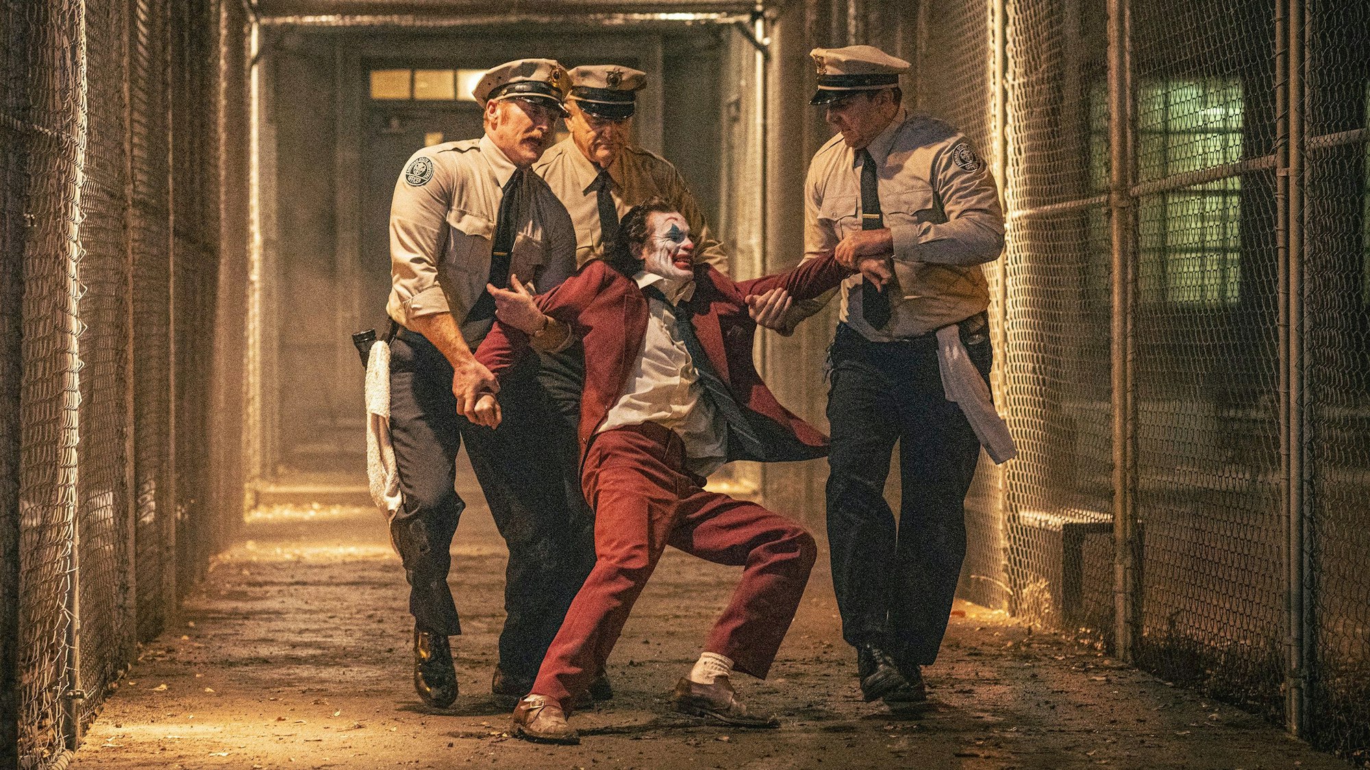 Drei Gefängnisaufseher schleppen einen sich heftig wehrenden Joaquin Phoenix im Joker-Kostüm durch einen Gang zwischen Maschendrahtzäunen zu seiner Zelle zurück.