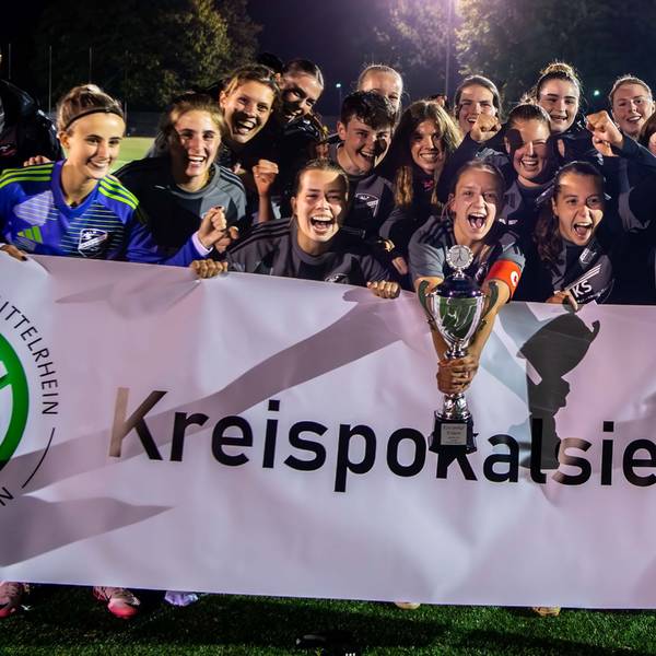 Hinter einem weißen Transparent mit der Aufschrift „Kreispokalsieger“ stehen die Spielerinnen des TB-SV Füssenich-Geich und jubeln. Die Kapitänin hält den silbernen Pokal in der Hand.