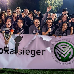 Hinter einem weißen Transparent mit der Aufschrift „Kreispokalsieger“ stehen die Spielerinnen des TB-SV Füssenich-Geich und jubeln. Die Kapitänin hält den silbernen Pokal in der Hand.
