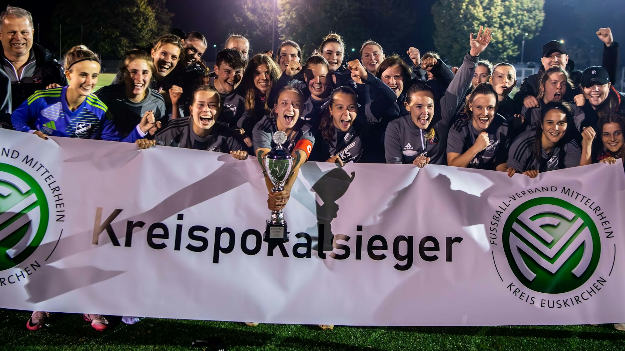 Hinter einem weißen Transparent mit der Aufschrift „Kreispokalsieger“ stehen die Spielerinnen des TB-SV Füssenich-Geich und jubeln. Die Kapitänin hält den silbernen Pokal in der Hand.