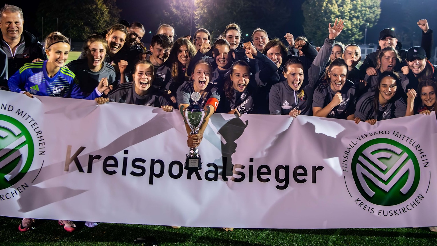 Hinter einem weißen Transparent mit der Aufschrift „Kreispokalsieger“ stehen die Spielerinnen des TB-SV Füssenich-Geich und jubeln. Die Kapitänin hält den silbernen Pokal in der Hand.