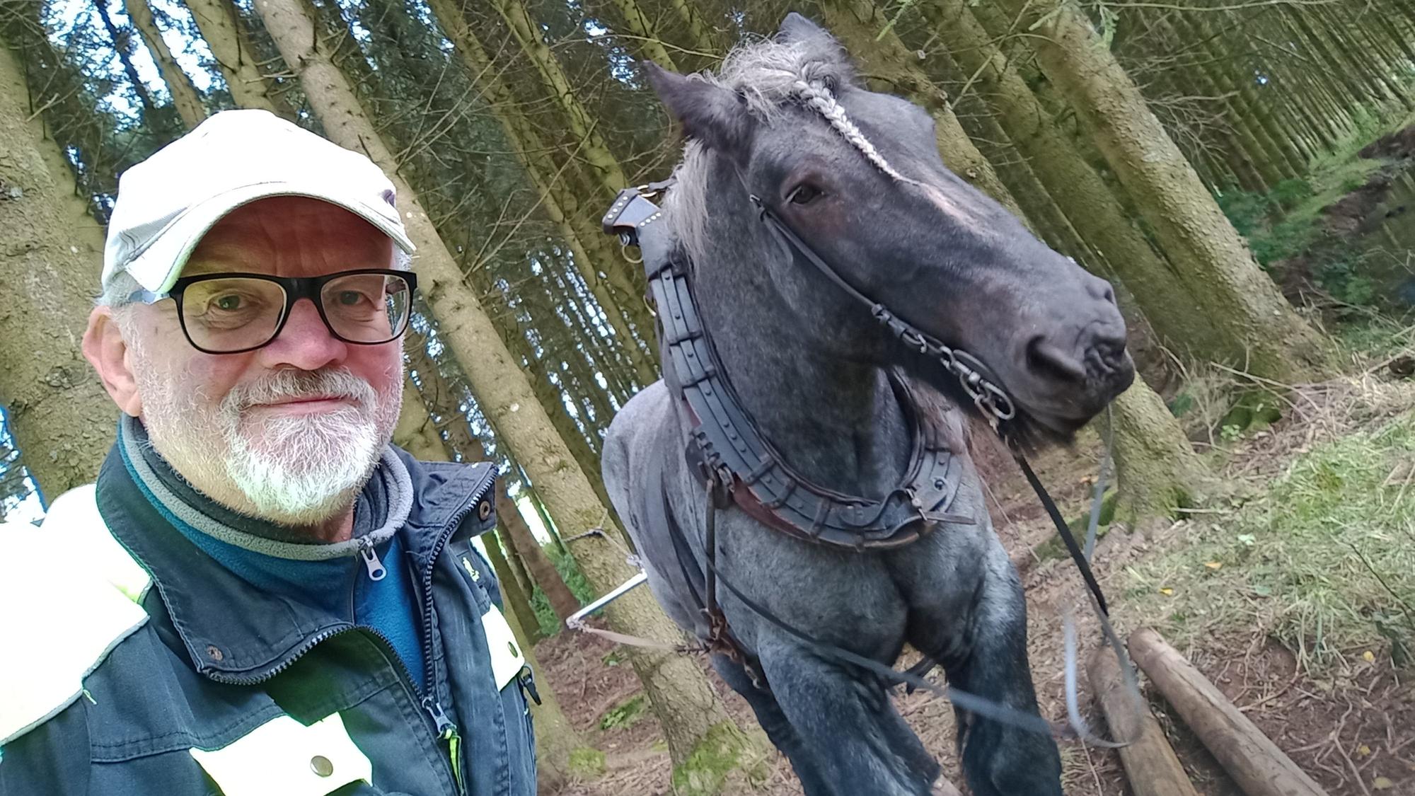 Ein Mann mit Brille und Kappe steht neben einem Pferd im Wald.