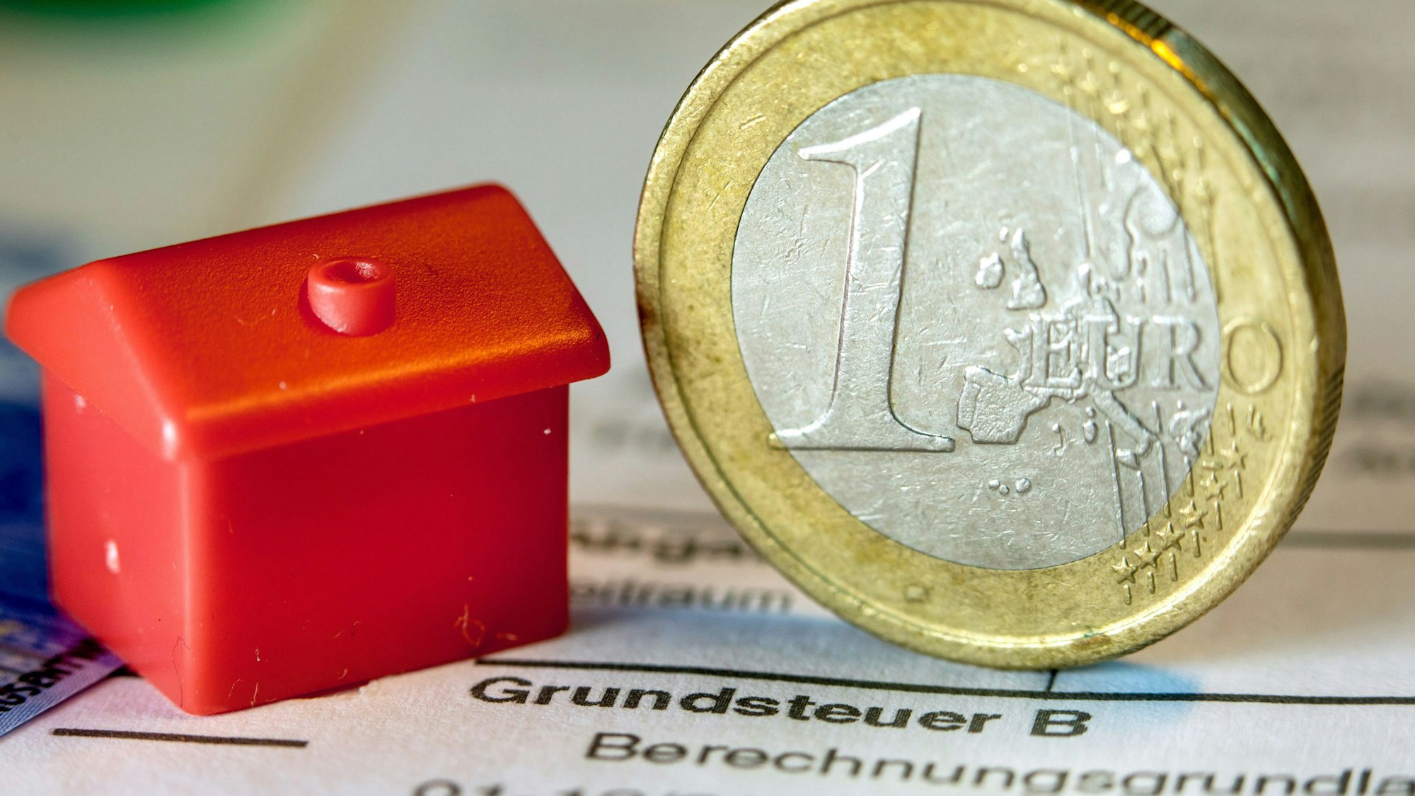 Eine Euro-Geldmünze sowie ein Spielzeughaus stehen auf einem Abgabenbescheid für die Entrichtung der Grundsteuer.