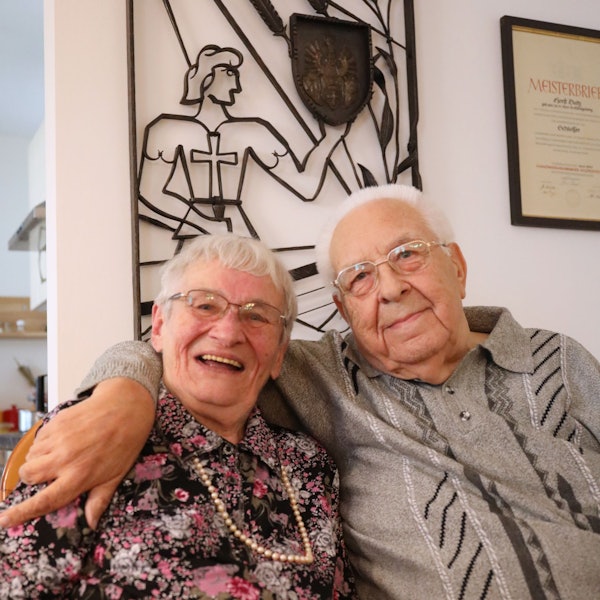 Ein Ehepaar sitzt eng beieinander, der Mann hat seinen Arm um seine Frau gelegt.