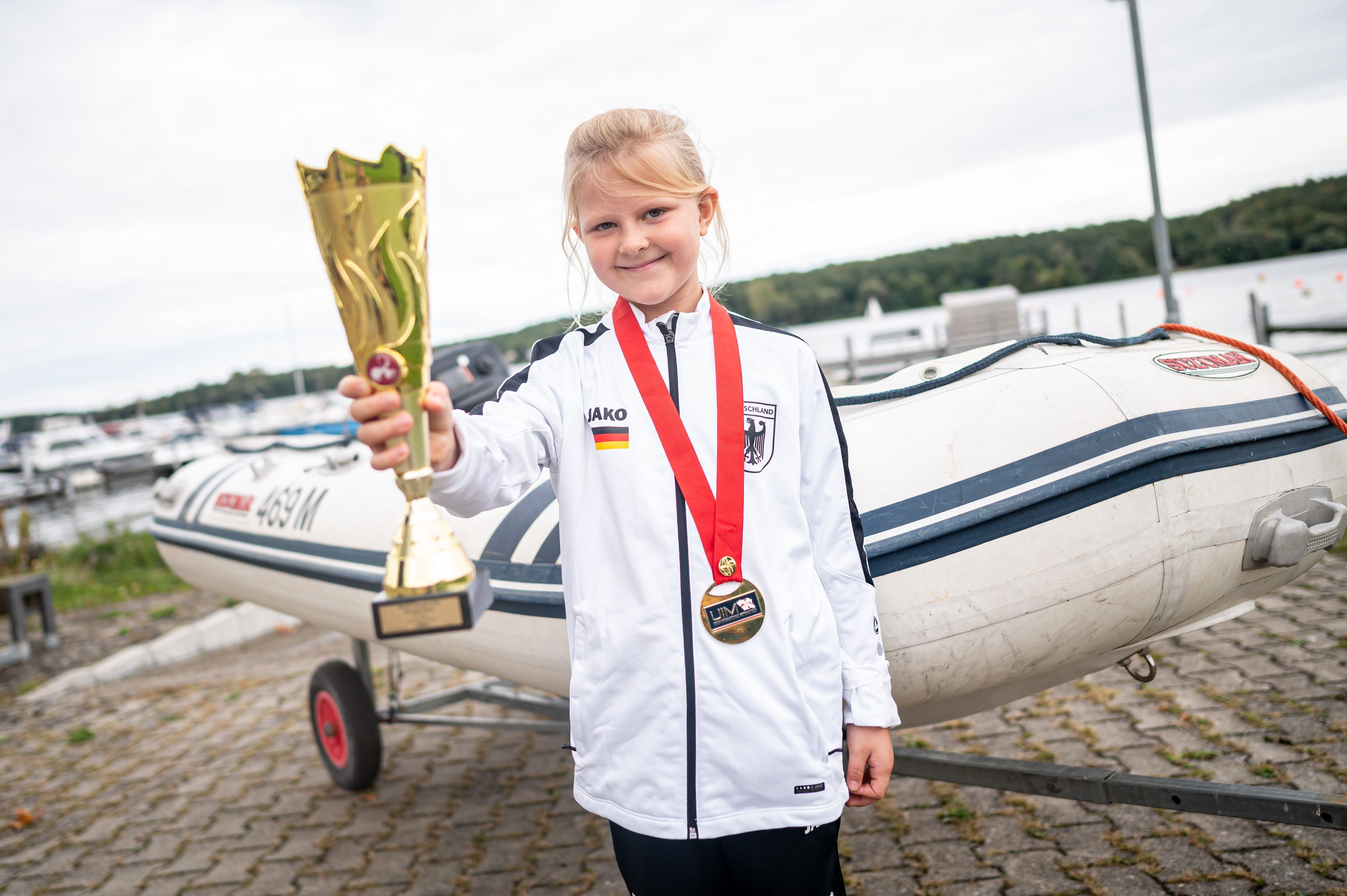 PRODUKTION - 25.09.2024, Brandenburg, Beetzsee/Brielow: Jana Pietack, siebenjährige Weltmeisterin im Schlauchbootfahren, steht mit Pokal und Medaille vor ihrem Schlauchboot. (zu dpa: «Siebenjährige Berlinerin ist Schlauchboot-Weltmeisterin») Foto: Fabian Sommer/dpa +++ dpa-Bildfunk +++
